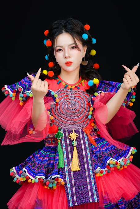 A woman in a vibrant pink and purple traditional ethnic costume adorned with colorful pom-poms and beadwork, posing with her fingers raised against a black background.