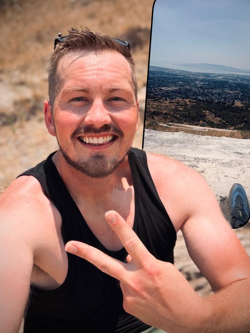 A smiling man in a black tank top flashes a peace sign while taking a selfie, with a scenic overlook featuring rolling hills, distant water, and clear blue skies visible in the background. The image aligns with the post's optimistic tone about enjoying nature and beautiful vistas, capturing a moment of genuine happiness in an elevated outdoor setting.