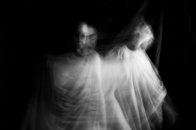 A black and white photograph captures two ghostly, blurred figures shrouded in translucent fabric against a dark background.