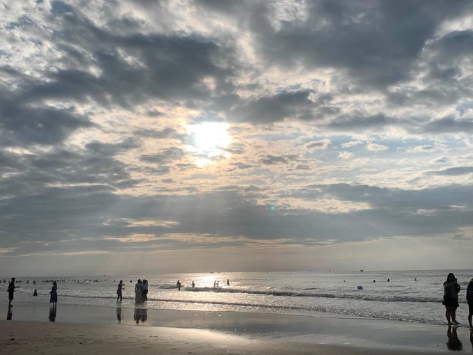 Beach scene at sunset or sunrise with sun breaking through cloudy skies, creating light rays over the ocean where small silhouettes of people are scattered along the shoreline.