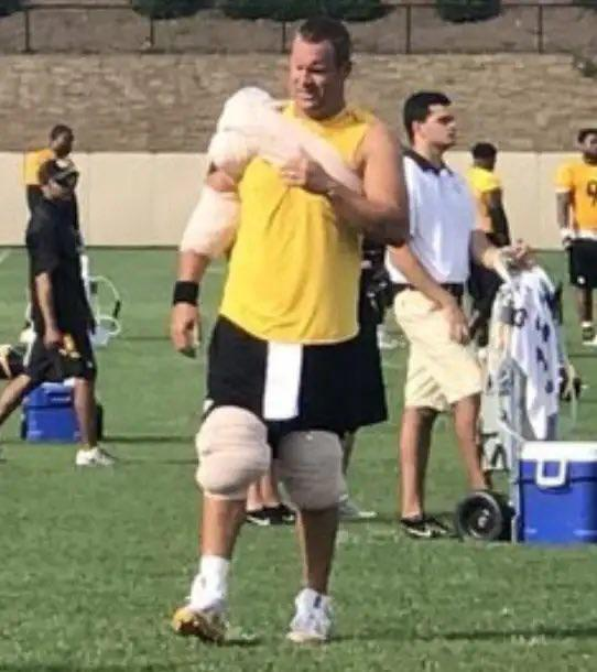 A photo from a football practice shows a heavyset player in a yellow sleeveless shirt and black shorts, wearing a sling/brace on his right arm and a knee brace on his left leg, standing on a field with a coach and other players nearby.