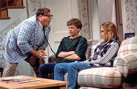 A comedic scene shows a doctor in a plaid jacket yelling with his mouth wide open at a young man and woman sitting on a couch.