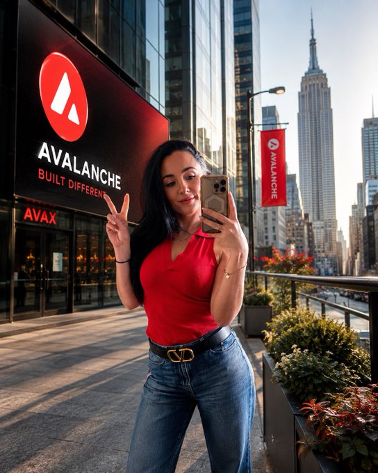 The image features a woman taking a selfie with her phone outside of an Avax building. The backdrop includes a city skyline, which suggests that they are located in New York City or another urban area with tall buildings. The focus of the photo is on the woman and her surroundings, capturing a moment from their daily lives.