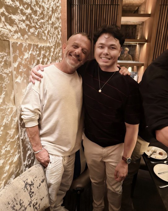 Two men smiling and posing together with their arms around each other at what appears to be a restaurant, with marble walls and dining tables visible in the background.