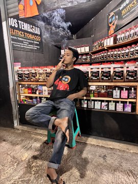 A young man wearing a black t-shirt, jeans, and a cap sits on a small green chair in a tobacco shop, smoking, with shelves of 'Elite's Tobacco' products behind him.