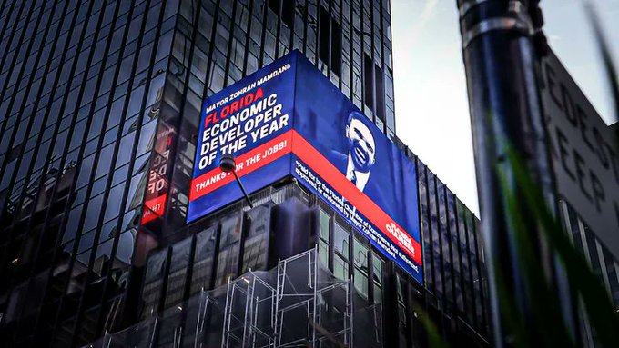 A billboard on a glass skyscraper reads "MAYOR ROKHAN MAMDANI FLORIDA ECONOMIC DEVELOPER OF THE YEAR THANKS FOR THE JOBS!" featuring a man's face.