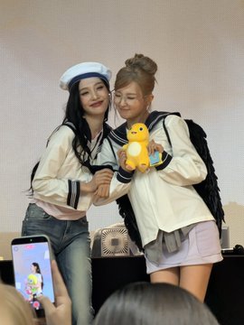 Two young women pose together at what appears to be a fan event, one wearing a sailor hat and outfit while the other holds a bright yellow Pokemon plush, with a smartphone visible in the foreground capturing the moment. The caption "JOYGI IS SO ALIVE" suggests the joyful, energetic atmosphere of the fan gathering, where the Pokémon plush (possibly Psyduck or Pikachu) and nautical-themed cosplay reflect the lively spirit of the community.