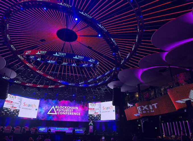 A large conference hall with a circular LED light installation on the ceiling and a stage displaying "BLOCKCHAIN FUTURIST CONFERENCE" along with "TEXIT" branding and a Search logo.