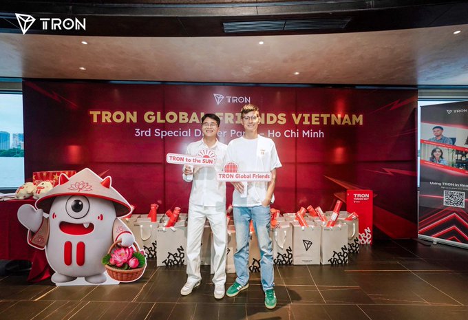 Two men pose in front of a red backdrop reading 'TRON GLOBAL FRIENDS VIETNAM 3rd Special Dinner Party Ho Chi Minh' at a TRON event, with one holding a 'TRON to the SUN' sign and another holding a 'TRON Global Friends' sign.