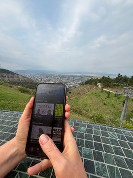 A person holds a smartphone displaying a trading app interface while standing outdoors overlooking a valley with a city in the distance, with a solar panel array visible beneath the phone.