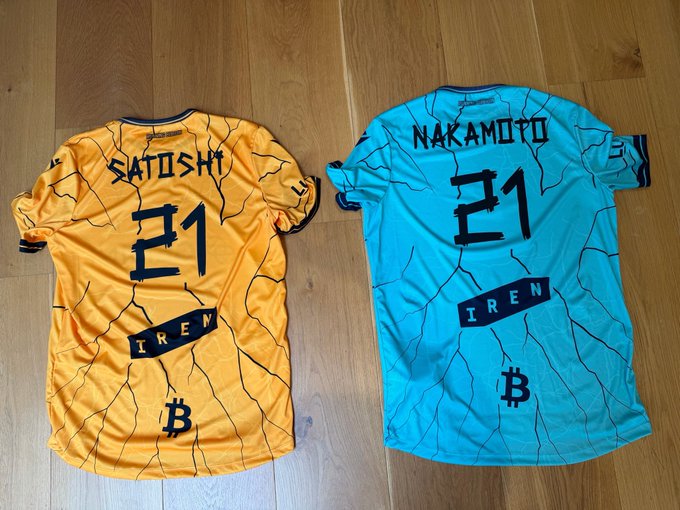 Two custom Bitcoin-themed soccer jerseys laid out on a wooden floor, designed to honor Satoshi Nakamoto, with 'SATOSHI 21' on the orange/gold jersey and 'NAKAMOTO 21' on the teal jersey, both featuring the 'IREN' logo and Bitcoin symbol.