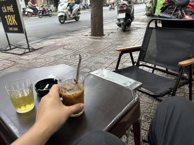 A first-person view of a hand holding an iced coffee at a small outdoor table with an ashtray and tea, situated on a sidewalk in Vietnam with a 'CÀ PHÊ 18K TAKE AWAY' sign and passing motorbikes in the background.