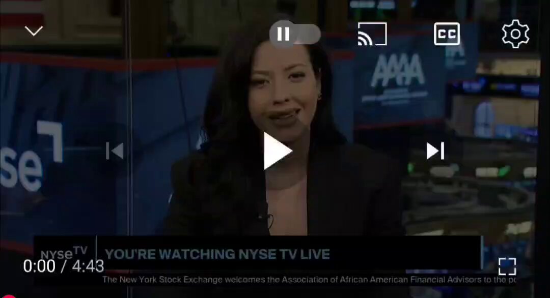 The image shows a woman sitting behind a desk, wearing a black blazer as she appears to be giving a presentation or speaking at an event. The background of the image features a TV screen displaying a video stream with text that reads "You're watching @nyse tv live". This suggests that the woman is likely presenting something related to the New York Stock Exchange (NYSE) on television, possibly discussing its history and significance in the financial world.