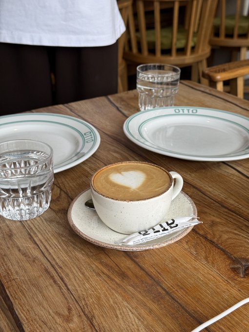 A cappuccino with heart-shaped latte art served in a ceramic cup on a saucer labeled 'OTTO', placed on a wooden table alongside two empty OTTO plates and glasses of water.