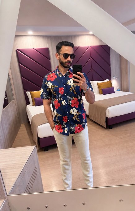The image features a man standing in front of two beds with white pillows, wearing a blue floral shirt and white pants. He is holding his phone up to take a selfie, capturing the moment. The room has purple walls and a wooden floor, creating an inviting atmosphere for the photo session.