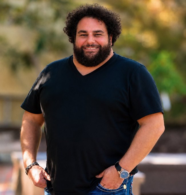 A portrait of a smiling bearded man with curly hair wearing a black V-neck t-shirt and a wristwatch, standing outdoors.