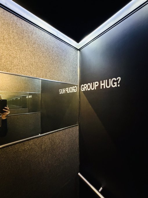 A dimly lit elevator interior with mirrored walls displaying the text 'GROUP HUG?' on the paneling, with a phone reflection visible.