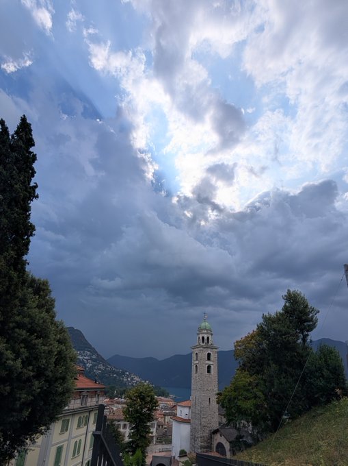 A dramatic sky filled with billowing gray clouds towers over a charming European town, where a tall stone bell tower with a green dome stands prominently among red-roofed buildings nestled at the foot of mountains. The moody, ominous atmosphere of the scene perfectly echoes the post's reference to a "Bitcoin apocalypse," with the dark, brooding clouds looming over Lugano like storm clouds over a financial downturn.