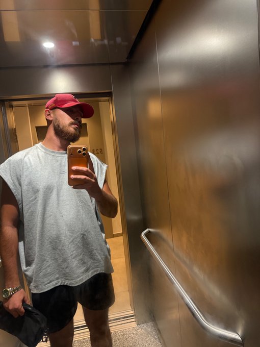 A bearded man wearing a red cap, gray sleeveless shirt, and dark shorts takes a mirror selfie with a gold iPhone inside an elevator. The casual "Gm friends" greeting matches his laid-back morning look captured in the elevator mirror.