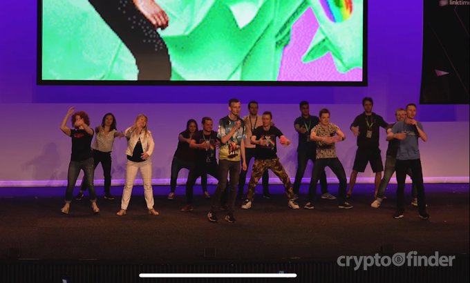 A group of people perform a synchronized dance on a stage in front of a large screen, watermarked with 'cryptofinder' in the bottom right corner.