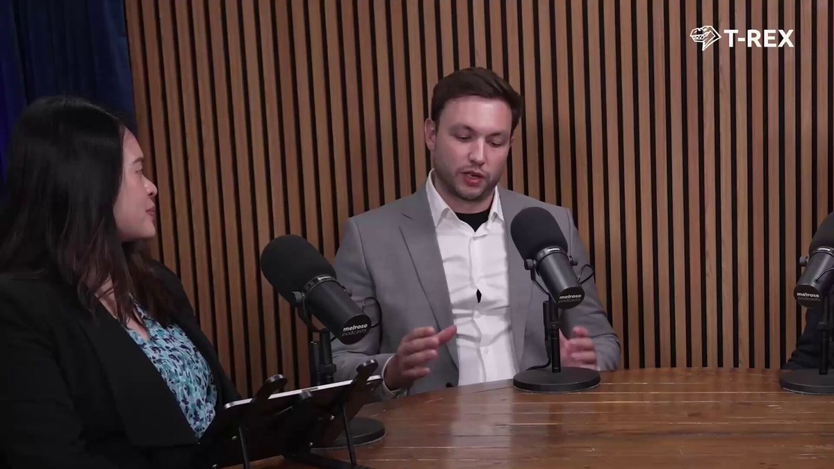 The image shows two people sitting at a table, one of them wearing a suit and the other a shirt. They are both holding microphones, suggesting that they might be participating in an interview or discussion. The background features wooden panels, giving the setting a professional and formal atmosphere. There is also text visible on the image, which reads "Tokenization without utility is just infrastructure The T-REX AppStore. ERC-3643 compatible applications that bring regulated assets to life. Issuance, distribution, yield, lending and more." This information might be related to a podcast or interview they are conducting, discussing the topic of tokenization in the context of the T-rex app store.