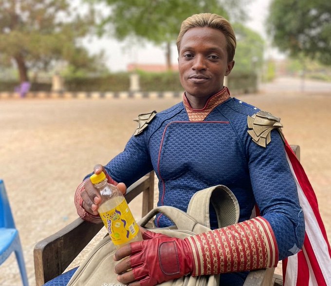 A man wearing a blue and red superhero costume with gold shoulder armor sits outdoors holding a bottle of orange "Teem" soda, with an American flag draped nearby.