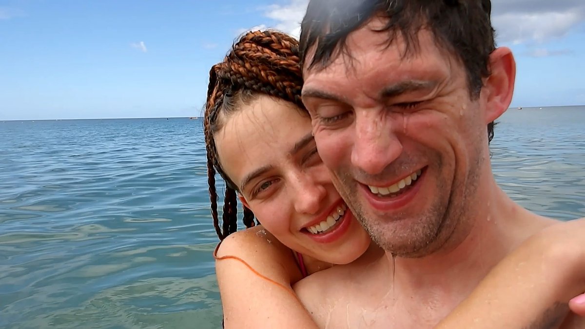 A selfie of a smiling couple in the ocean, with the woman having braided hair resting against the man, both squinting in the bright sunlight.