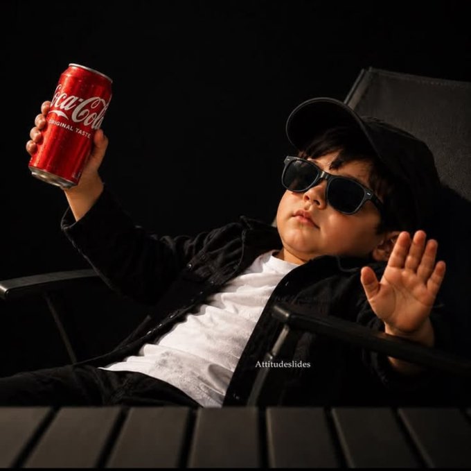 A young child wearing sunglasses, a black cap, and a black jacket over a white shirt, sitting in a chair and holding up a can of Coca-Cola Original Taste against a dark background.