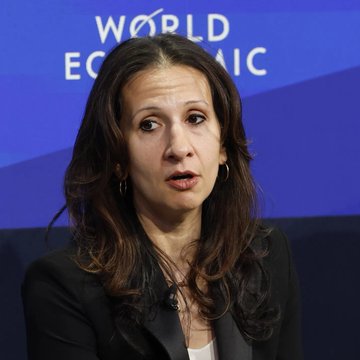 A woman with dark hair wearing a black blazer speaks at a World Economic Forum event, indicated by the blue backdrop behind her.