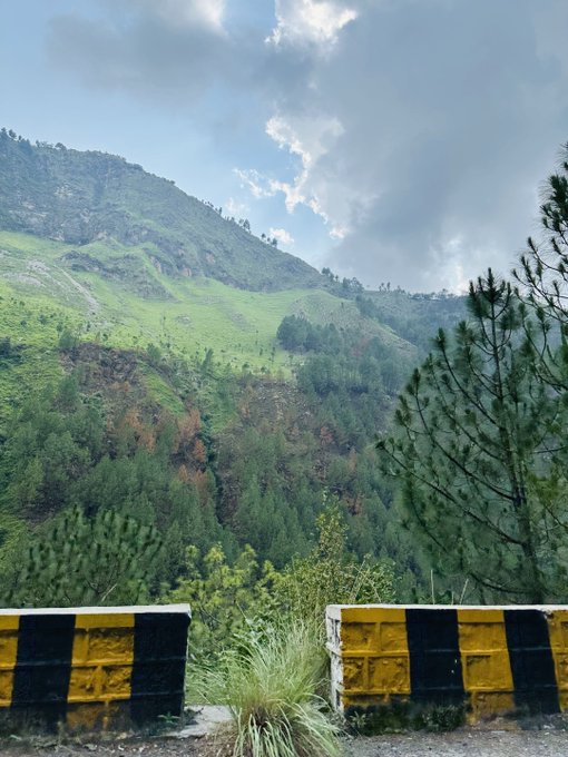 A scenic mountain landscape with lush green slopes, pine trees, and a partly cloudy sky, framed by yellow and black striped barriers in the foreground.