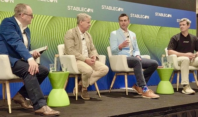 A panel discussion takes place at StableCon, with four men seated on stage against a green and blue branded backdrop reading "STABLECON." The central speaker gestures animatedly while engaging in debate with his fellow panelists, capturing the lively exchange described in the post about discussing the future of finance.