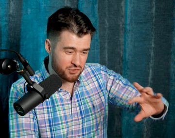 A man with dark hair and a beard, wearing a blue and pink plaid shirt, speaks animatedly into a black studio microphone while gesturing with his hand, set against a teal velvet curtain backdrop—capturing the expressive, skeptical mimicry of crypto trading enthusiasts referenced in the caption.