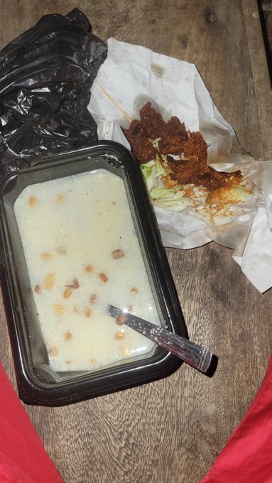 A creamy soup in a black takeout container with a spoon sits beside a piece of dark, seasoned fried meat on paper and a crumpled black plastic bag, all resting on a wooden table. The meal appears to be a takeout spread ready to be enjoyed, fitting the post's casual mention of using the food to hold over for the night.