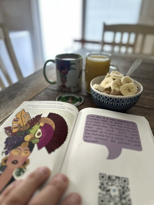 A cozy breakfast scene featuring a mug of coffee, a glass of orange juice, and a bowl of cereal topped with banana slices next to an open book with colorful illustrations and a QR code.