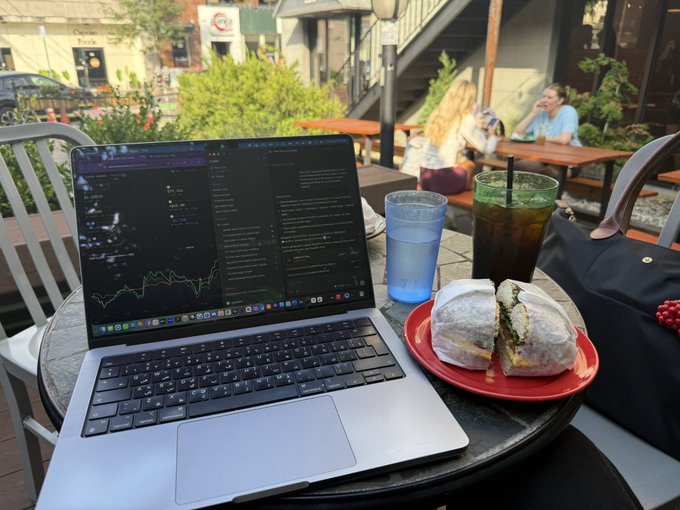 An outdoor cafe scene with a MacBook displaying a financial trading chart, accompanied by a bagel sandwich on a red plate and two beverages (water and iced drink) on a metal table.