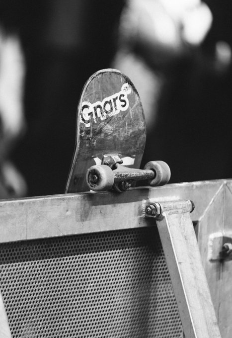 A black and white photograph of a worn skateboard with 'Chriss' written on it, perched on the edge of a metal ramp with mesh grating.