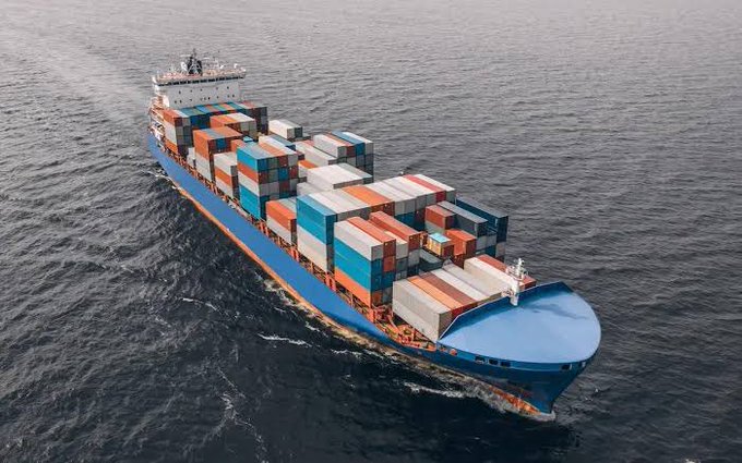 An aerial photograph shows a large blue container ship loaded with multi-colored shipping containers sailing through dark blue ocean waters.