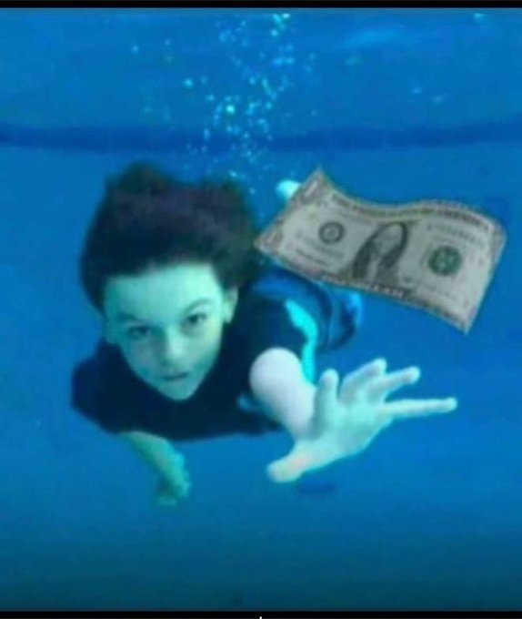 An underwater photograph of a young boy swimming while holding a one dollar bill, reaching toward the camera in a pool.
