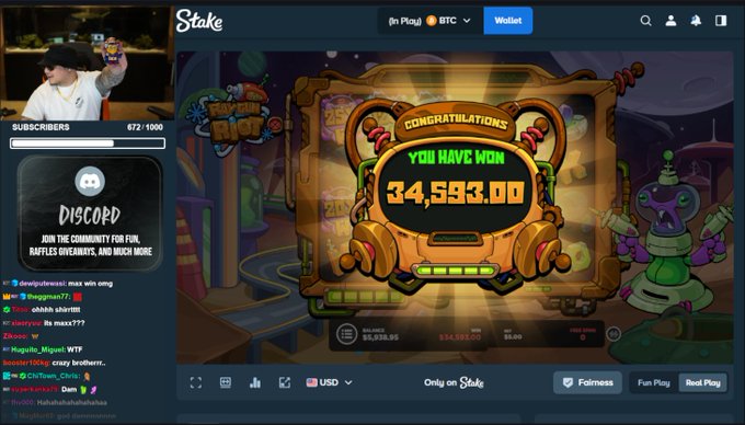 A streamer celebrates a massive $34,593.00 win on the slot game "Raygun Riot" at Stake casino, showing the "CONGRATULATIONS YOU HAVE WON" screen with betting details of a $5.00 wager and $34,593.00 win. The stream chat and viewer count (672/1000) are visible alongside a Discord community promotion banner, matching the post's excitement over hitting a huge payout on a small bet.