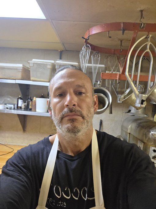 A man with a graying beard wearing a black shirt and apron takes a selfie in a kitchen, with whisks and other baking equipment hanging from an orange rack behind him.