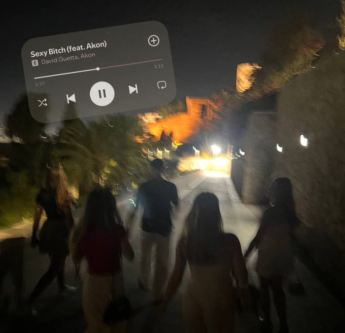A blurry night photograph of a group of people walking along an illuminated outdoor path, overlaid with an Apple Music player interface showing the song 'Sexy Bitch (feat. Akon)' by David Guetta, Akon.