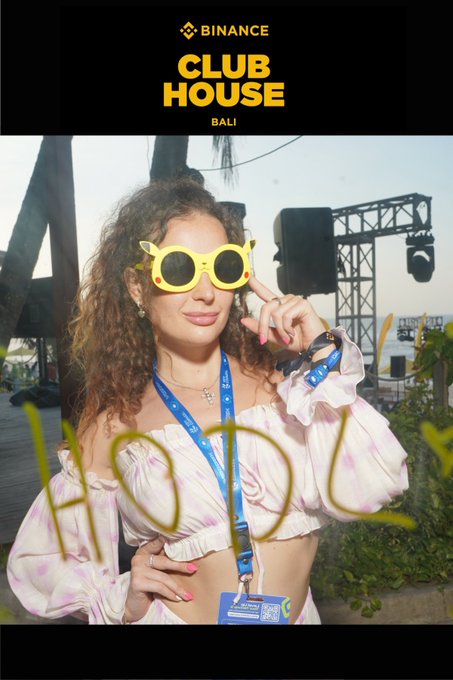A woman with curly hair wearing yellow sunglasses and a purple off-shoulder top poses at the Binance Club House in Bali, with 'HOLY' written in the foreground.