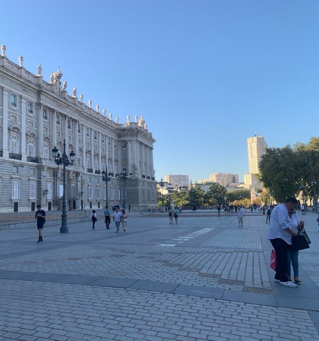 A grand plaza with classical architecture, ornate lampposts, and pedestrians enjoying a clear sunny day, evoking a cheerful "gm" (good morning) vibe with its bright, welcoming atmosphere. The scene shows people walking and gathering near a stately historic building, perfect for a positive morning greeting.