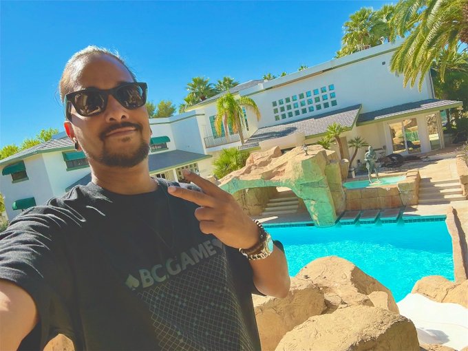 A man wearing sunglasses and a "BC.GAMES" shirt takes a selfie in front of a luxurious villa with a pool, palm trees, and rocky waterfall feature, embodying a relaxed, aspirational "Gm" lifestyle vibe.