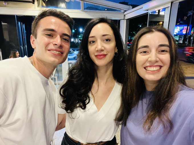 A selfie of three smiling young adults—two women and one man—taken indoors at night near a window.
