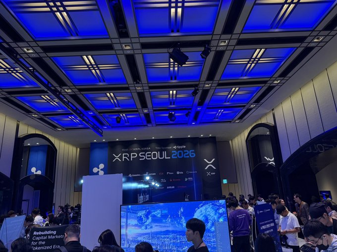 A large conference hall with a blue-lit ceiling hosts the 'XRP SEOUL 2026' event, featuring the XRP/Ripple logo and numerous attendees gathered in the venue.