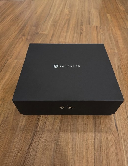 A black box with the 'TOKENLON' logo on top and '7th' written on the side, sitting on a wooden floor.