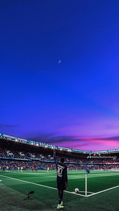 A soccer player stands on the field at night with a crescent moon visible in the blue sky above the stadium.