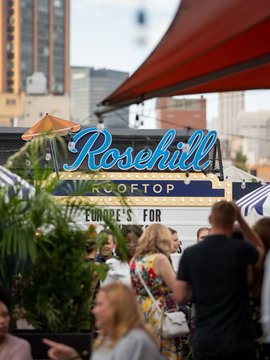 A rooftop bar scene featuring a neon sign reading "Rosehill ROOFTOP" with a marquee below partially displaying text about Europe, while a crowd of people mingle underneath in what appears to be a premier social gathering spot — perfectly matching the post's invitation to a legendary crypto networking event with @fomo and @privy_io.