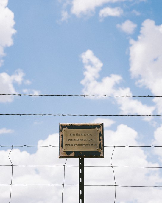 A weathered plaque mounted on a metal post in front of a wire fence and blue sky with clouds, reading 'Blue Sky #13, 2025, Daniel Koeth (b. 1993), Owned by Benny Red Beard'.